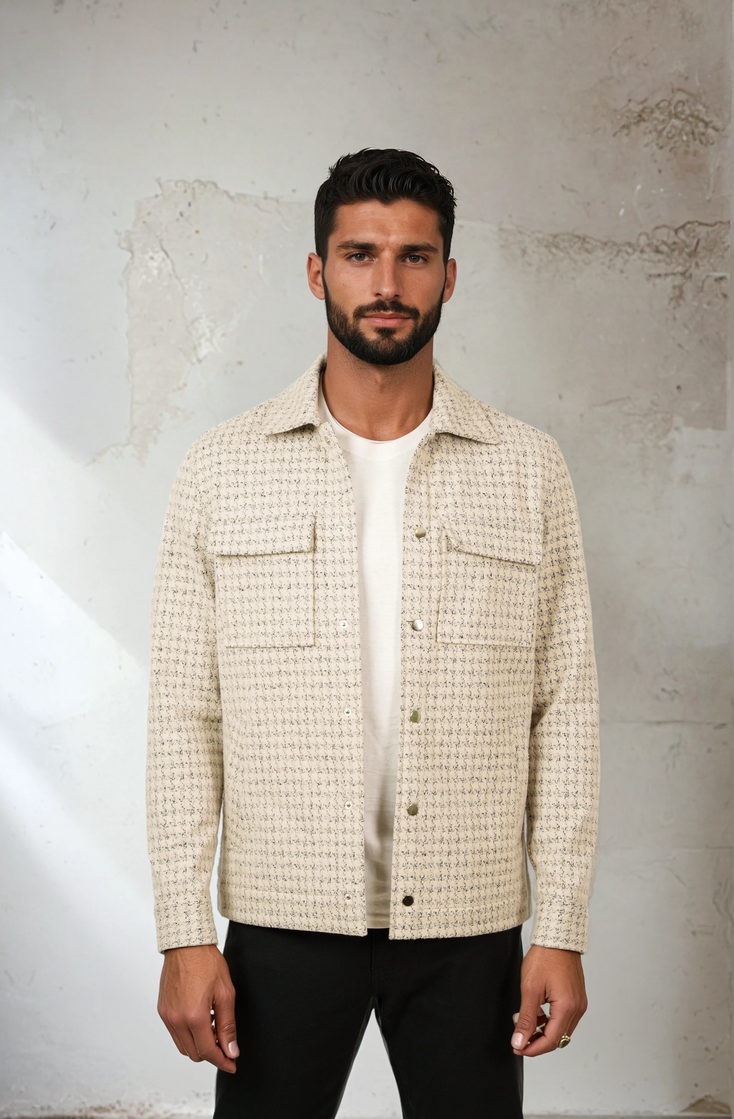 Man wearing a checkered shirt over a white t-shirt against a textured wall.