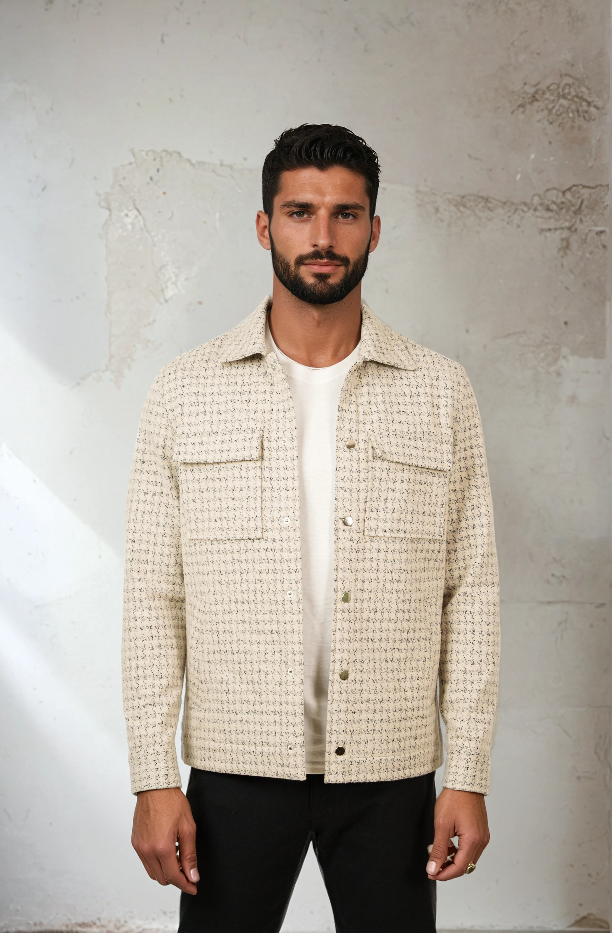 Man wearing a checkered shirt over a white t-shirt against a textured wall.