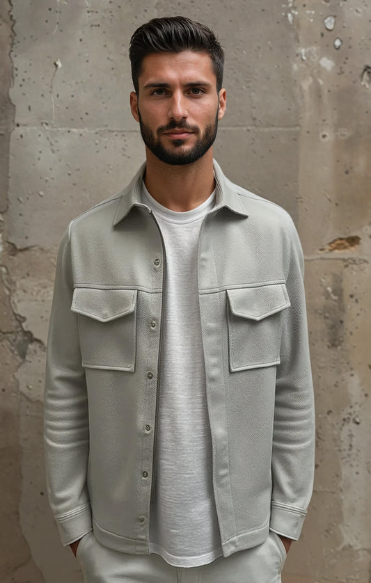 Man wearing a light gray shirt over a white t-shirt against a textured wall.