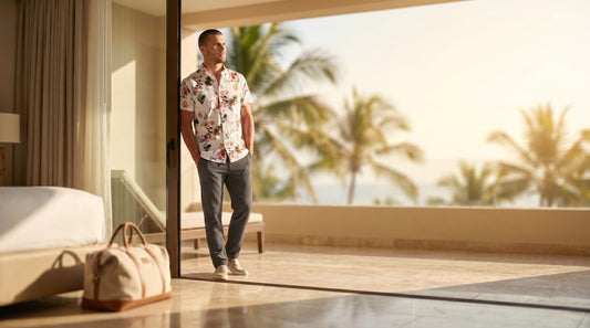 A man in a Stone Rose floral print short-sleeve shirt stands in the doorway of a sunlit resort hotel room, a canvas weekender bag on the floor beside the bed, palm trees and ocean beyond the terrace.