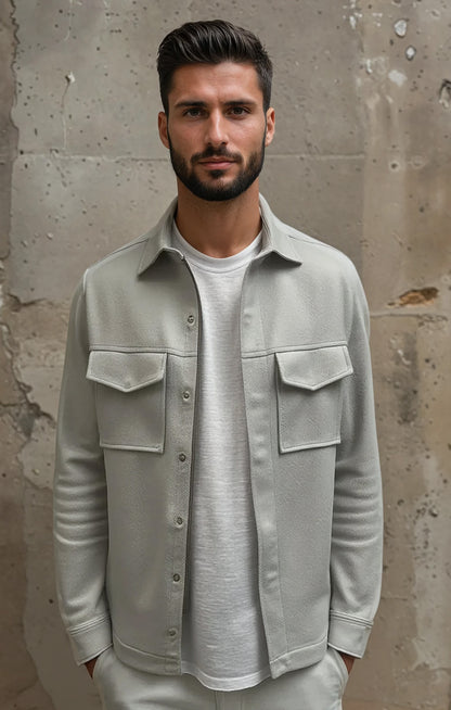 Man wearing a light gray shirt over a white t-shirt against a textured wall.